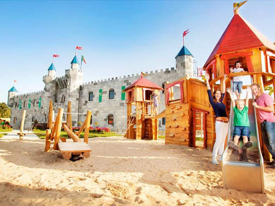 Een grote houten speeltuin met klimtoestellen en zand, direct naast het imposante LEGO kasteelhotel van LEGOLAND Duitsland.