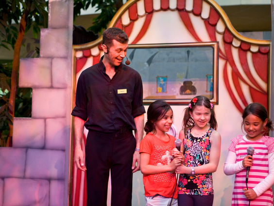 Drie meisjes staan samen met een entertainer lachend op het podium tijdens een kindershow in Center Parcs Limburgse Peel.