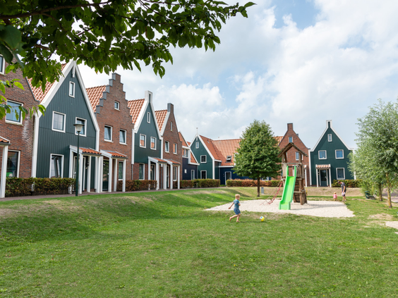 Een speeltuintje met glijbaan op een groot grasveld, omringd door karakteristieke vakantiehuizen in Volendamse stijl