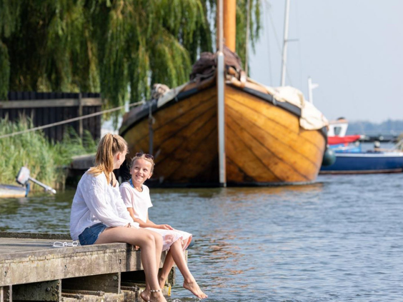 Een gezin zit met de voeten boven het water op een steiger in de haven, met een klassiek houten schip op de achtergrond.