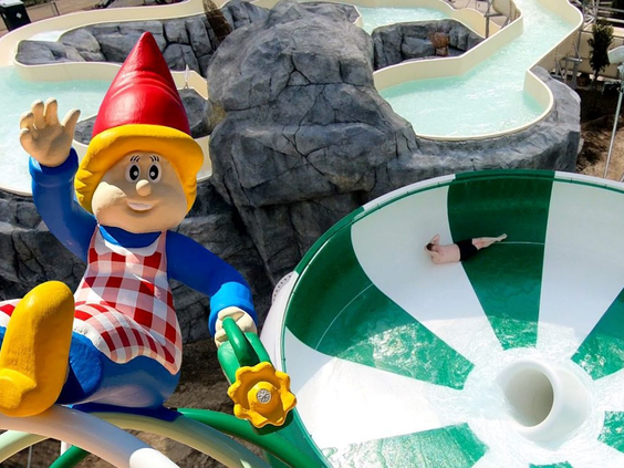 Een vrolijke kabouter mascotte van Marveld Recreatie staat bij de grote groen-witte trechterglijbaan in het waterpark.