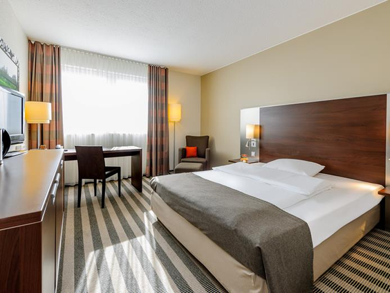 Overzicht van een ruime en moderne hotelkamer in het Mercure Duisburg City, met een groot tweepersoonsbed en gestreepte vloerbedekking.