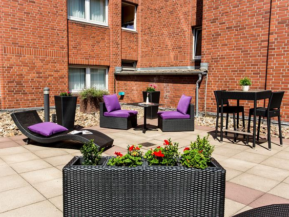 Het gezellige buitenterras van het Mercure Hotel in Duisburg City met comfortabele zitjes met paarse kussens, perfect om buiten te zitten.