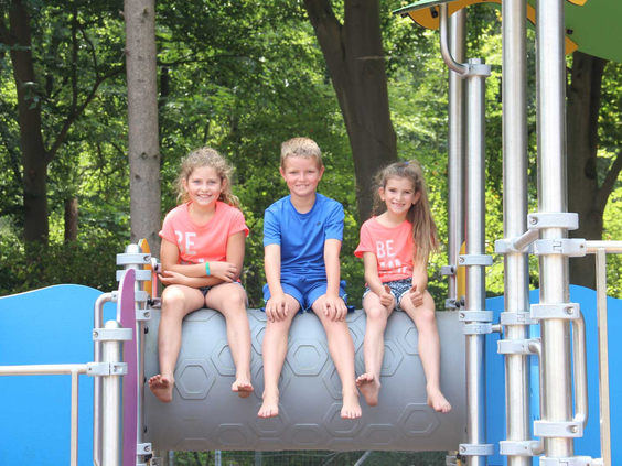 Drie kinderen, een jongen en twee meisjes, zitten lachend naast elkaar op een speeltoestel in de speeltuin van Bosbad Hoeven.