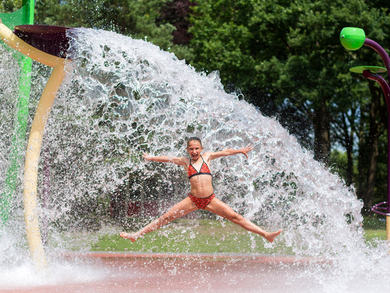 Een meisje in bikini springt vrolijk met gespreide armen en benen onder een groot watergordijn in het spray-park van Waterspeelpark Splesj.