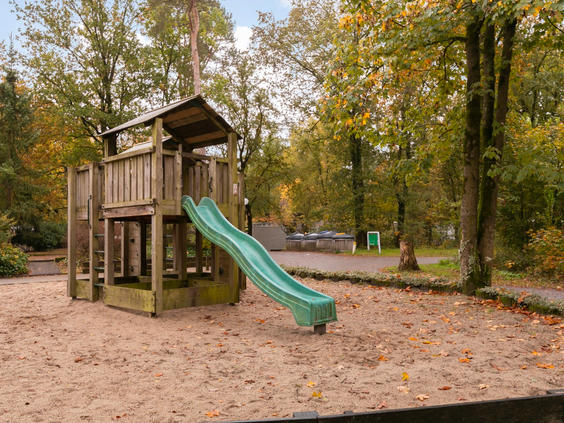 Een houten speeltoestel met glijbaan in een zandbak, gesitueerd in de bosrijke omgeving van Parc de Berkenhorst.