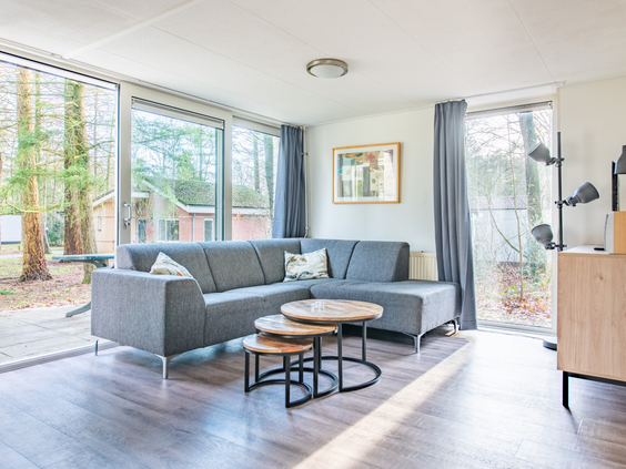 Lichte, moderne woonkamer met een grijze hoekbank en grote ramen die uitkijken op het bos bij Parc de Berkenhorst.