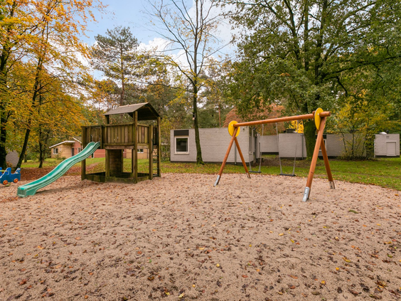 Een speeltuin met een grote zandbak, een glijbaan en schommels, gelegen tussen de bomen op Parc de Berkenhorst.