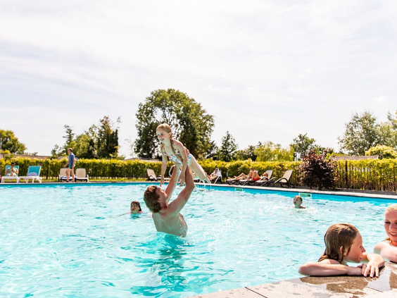 Een vader gooit zijn kind lachend in de lucht in het verwarmde buitenzwembad van Parc de IJsselhoeve, vol waterpret.
