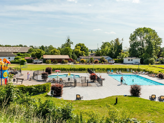 Overzicht van het zonnige buitenzwembad met een gezellig terras en de grote speeltuin op de achtergrond.