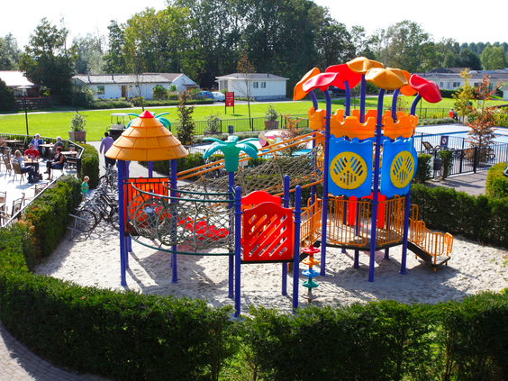 Overzicht van de omheinde en ruime buitenspeeltuin met een groot, kleurrijk speeltoestel en veel zand om in te spelen.