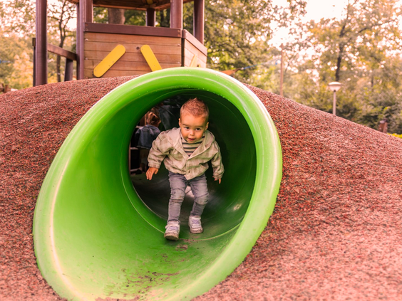 Een jong kind kruipt lachend uit een groene tunnelglijbaan in een van de speeltuinen van Parc Heihaas.