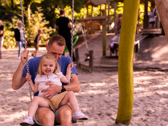 Een vader en zijn lachende dochter zitten samen op de schommel in de speeltuin van Parc Heihaas.