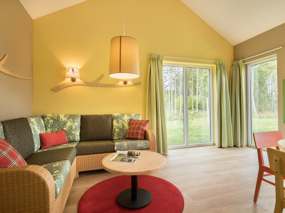 De gezellige woonkamer van een Premium cottage in Park Allgäu, met een comfortabele hoekbank, kleurrijke details en openslaande deuren naar het terras en het bos.