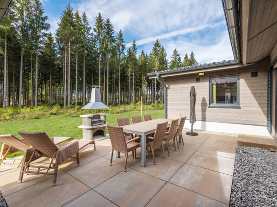 Het ruime privéterras van een Exclusive cottage in Park Allgäu, uitgerust met een grote eettafel, ligbedden en een stenen barbecue, direct aan de rand van het bos.