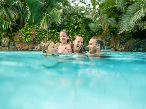 Een lachend gezin met twee kinderen ontspant samen in het helderblauwe water van het tropische zwembad van Aqua Mundo in Center Parcs Park Allgäu.
