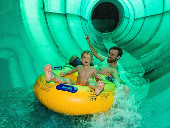 Een gezin met een kind glijdt lachend in een grote gele zwemband door een groene, verlichte bandenglijbaan in waterpark Aqua Mundo van Park Allgäu.