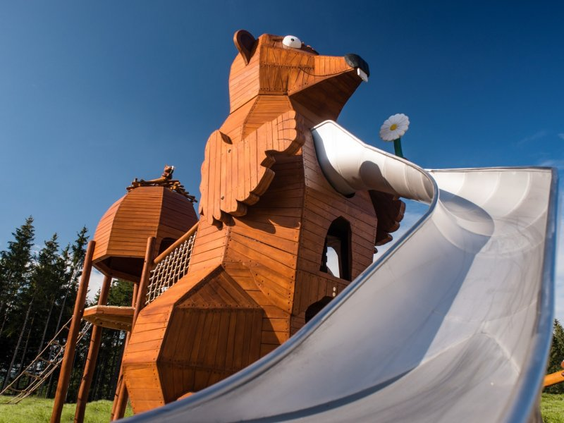 Een groot houten speeltoestel in de vorm van een beer met een lange metalen glijbaan in de buitenspeeltuin van Center Parcs Park Allgäu.