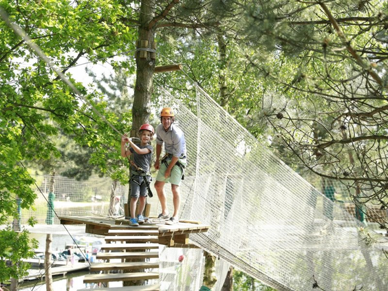 Een vader en zoon met veiligheidshelmen en harnassen op een hoog touwenparcours tussen de bomen in het klimpark van Center Parcs Park Allgäu.