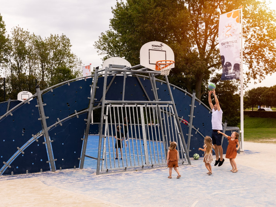 Kinderen en een volwassene spelen basketbal en voetbal in de multifunctionele sportkooi in het zand op Vakantiepark Port Greve.Kinderen en een volwassene spelen basketbal en voetbal in de multifunctionele sportkooi in het zand op Vakantiepark Port Greve.