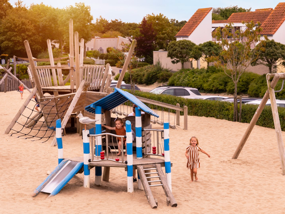 Twee jonge kinderen spelen in een grote zandspeeltuin met een houten klimtoestel en glijbaan op Vakantiepark Port Greve.
