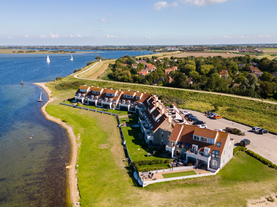 Een prachtige luchtfoto van Vakantiepark Port Greve aan het water in Zeeland, met een rij vakantiehuizen langs de kustlijn en zeilboten op het water.