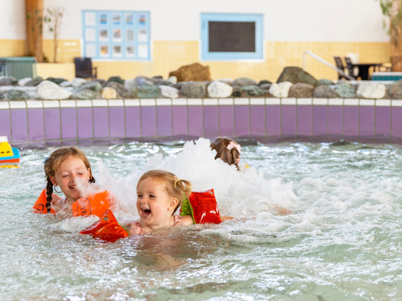 Twee lachende meisjes met zwembandjes spelen en spetteren in het ondiepe, bruisende kinderbad van het zwembad op Vakantiepark Port Greve.