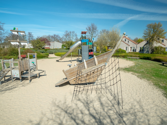 Een grote buitenspeeltuin in het zand op Vakantiepark Port Greve, met een houten piratenschip om op te klimmen en een glijbaan.