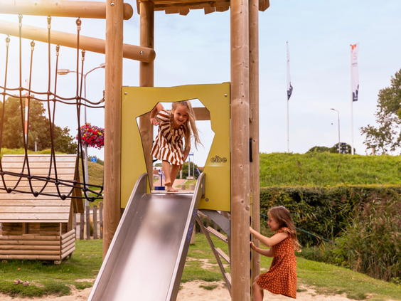 Twee meisjes spelen in de buitenspeeltuin van Vakantiepark Port Greve, een meisje glijdt van de glijbaan terwijl de ander toekijkt.
