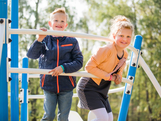 Een broer en zus lachen naar de camera terwijl ze samen op een blauw klimrek spelen in de speeltuin van Recreatiepark Beekbergen.