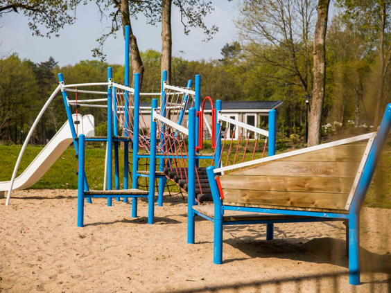 Overzicht van de buitenspeeltuin met zandondergrond, een glijbaan en een groot blauw klimtoestel op Recreatiepark Beekbergen.