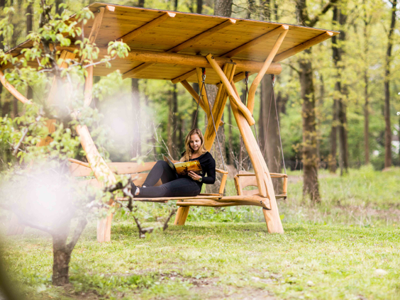 Een gast ontspant met een boek op een houten schommelbank midden in de bossen van Recreatiepark Beekbergen.
