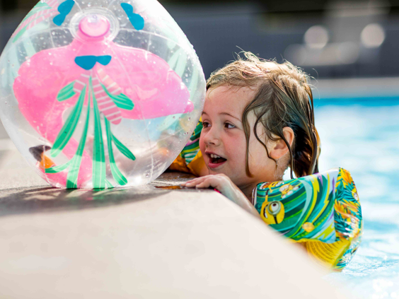 Lachend meisje met zwembandjes leunt op de rand van het zwembad en houdt een vrolijke strandbal vast op Recreatiepark Beekbergen.
