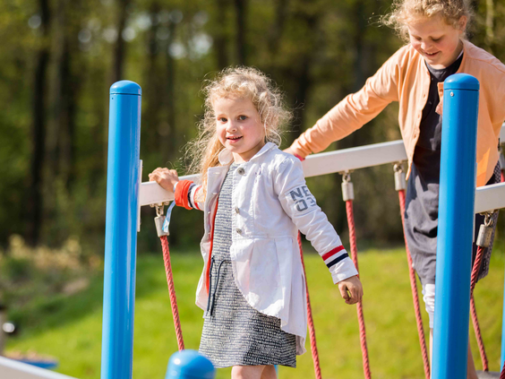 Een meisje met blonde krullen balanceert lachend over een wiebelige touwbrug in de buitenspeeltuin van Recreatiepark Beekbergen.
