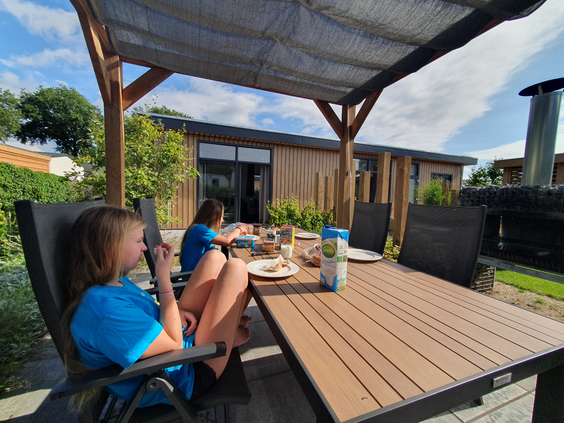 Een gezin geniet samen van het ontbijt aan een grote tafel op het overdekte, houten terras van hun vakantiehuis op Recreatiepark de Boshoek.