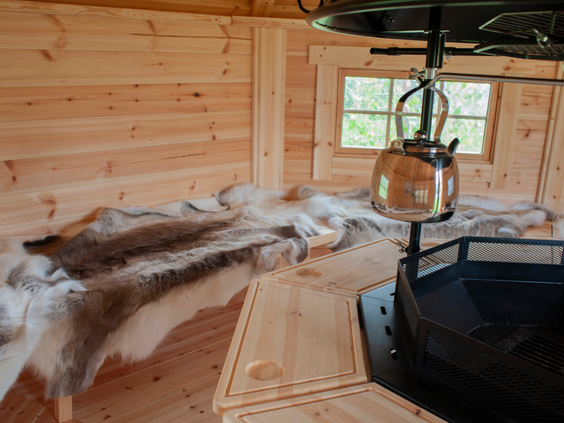 Het houten interieur van een Finse Grillkota op Recreatiepark de Boshoek, met een centrale grill en houten banken met dierenvellen.