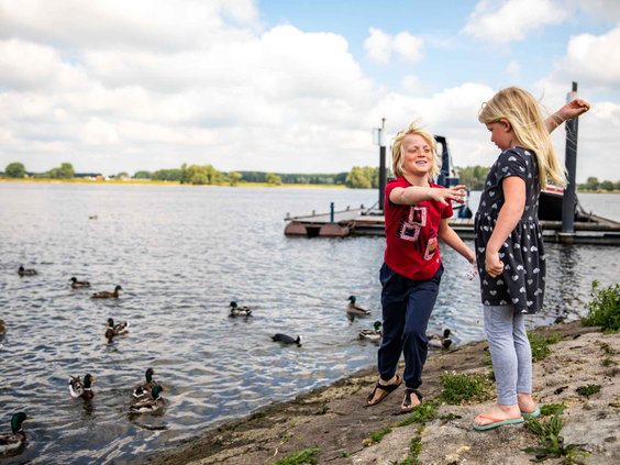 Twee kinderen staan aan de waterkant en voeren de vele eendjes die zwemmen in recreatieplas het Esmeer.