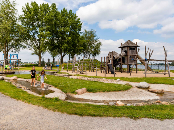 Kinderen spelen in de natuurlijke waterspeeltuin met een waterloop en een groot houten speelkasteel op de achtergrond.