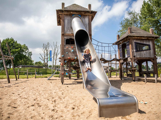 Een meisje glijdt van de hoge metalen glijbaan van het houten speelkasteel in de zandspeeltuin van het recreatiepark.