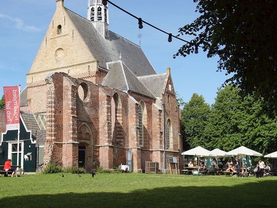 De historische Ruïnekerk in het centrum van het kunstenaarsdorp Bergen, omringd door groen en een gezellig terras.