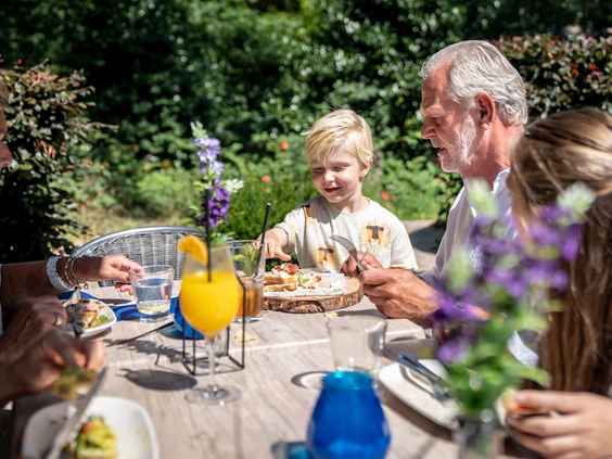 Een lachende familie met kinderen geniet van een lunch buiten aan een houten tafel in de tuin van hun vakantiehuis.