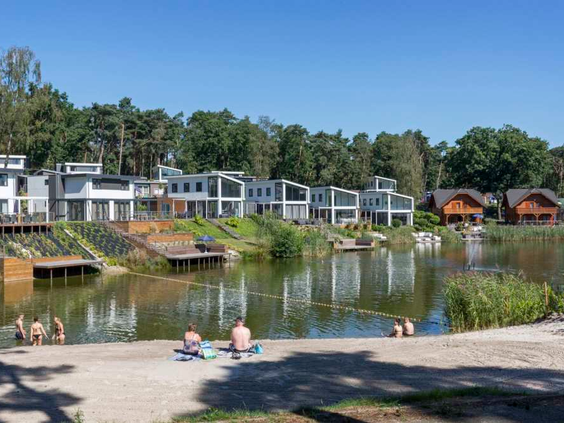 Gasten zwemmen en zonnen bij de recreatieplas met zandstrand op Resort Brunssummerheide in Limburg, met uitzicht op de luxe waterwoningen.