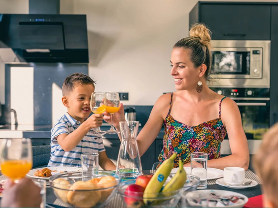 Een lachende moeder en zoon proosten met een glas jus d'orange aan een rijkelijk gevulde ontbijttafel.