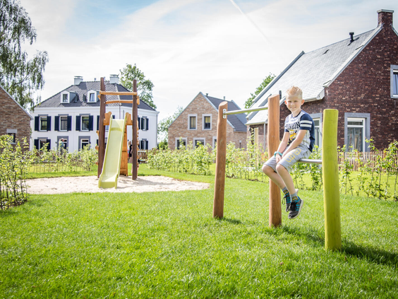 Een jongen speelt op een speeltoestel in een groene, parkachtige speeltuin tussen de vakantiehuizen van Dormio Resort Maastricht.