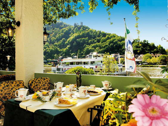 Het terras van Hotel Bellevue met een gedekte tafel en uitzicht op de kasteelruïne Grevenburg en de wijngaarden van de Moezel.