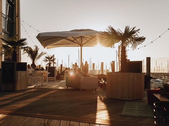 Het sfeervolle houten terras van de beachclub op Roompot Beach Resort met loungebanken en uitzicht op de jachthaven tijdens zonsondergang.
