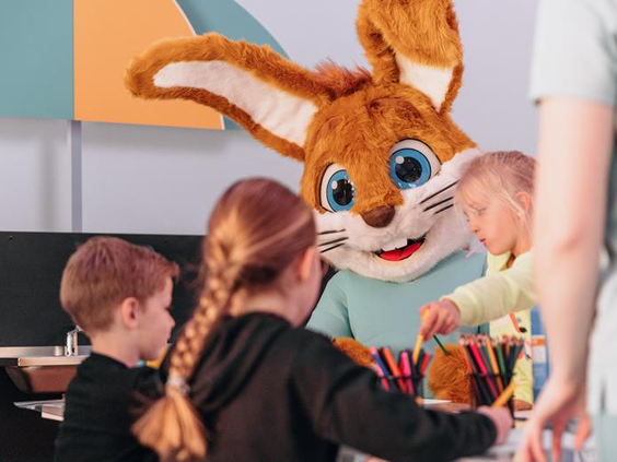 Mascotte Koos Konijn knutselt gezellig samen met een groepje kinderen aan een tafel in de Koos Kids Club.