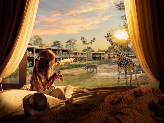 Een meisje met een knuffelgiraffe in haar arm wijst vanuit haar lodge enthousiast naar de echte giraffen buiten bij safari hotel beekse bergen