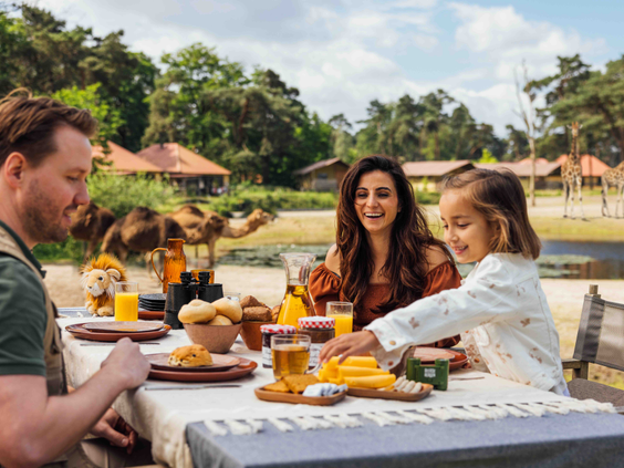 Gezin met kind ontbijt op het terras van hun vakantiehuisje met uitzicht op kamelen en giraffen op Safari Resort Beekse Bergen