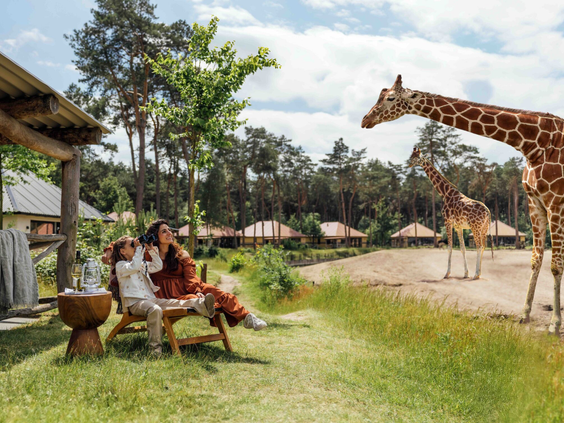 Moeder en dochter genieten op het terras van hun luxe lodge van het uitzicht op een giraffe bij Safari Resort Beekse Bergen